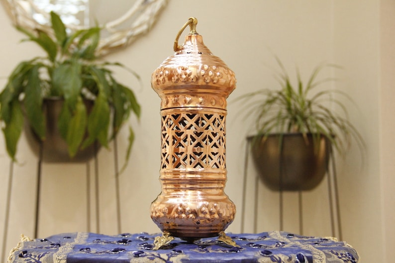 May include: A copper-colored lantern with an ornate design. The lantern has a handle at the top and sits on three small feet. The middle section features a lattice pattern. The lantern is placed on a blue patterned cloth.