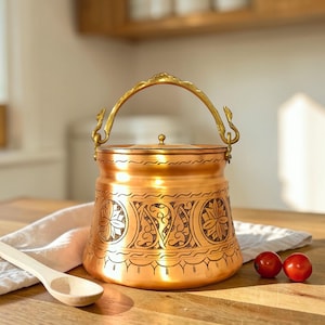 May include: A copper-colored pot with a decorative handle and lid. The pot features intricate etched designs around the body. A wooden spoon and two red tomatoes are on the wooden surface.