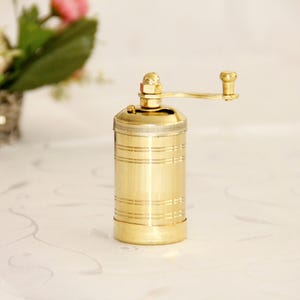 May include: A gold metal pepper grinder with a hand crank. The grinder has a ribbed design and is sitting on a white surface.