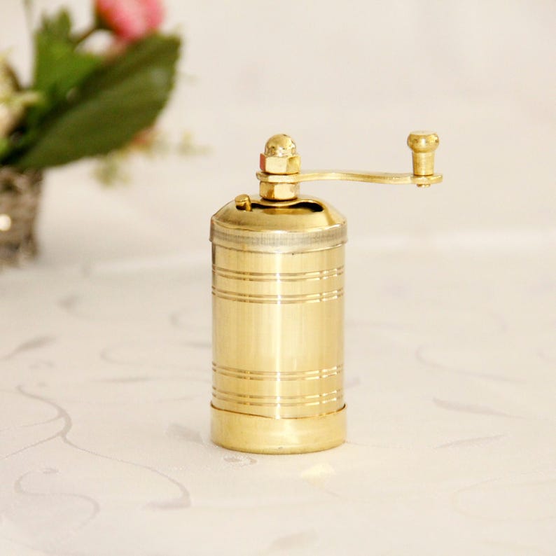 May include: A gold metal pepper grinder with a hand crank. The grinder has a ribbed design and is sitting on a white surface.