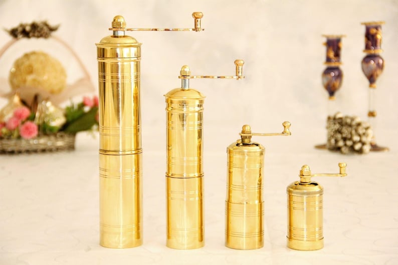 May include: Four gold-colored manual pepper grinders of varying sizes. The grinders have a cylindrical shape with a crank on the side.