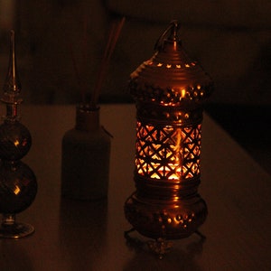 May include: A warm-toned image features a decorative lantern with intricate cut-out patterns, emitting a soft, inviting glow. Beside it are a glass decorative object and a small bottle. The scene evokes a sense of cozy ambiance.