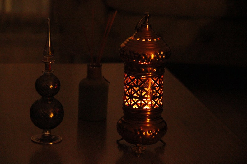 May include: A warm-toned image features a decorative lantern with intricate cut-out patterns, emitting a soft, inviting glow. Beside it are a glass decorative object and a small bottle. The scene evokes a sense of cozy ambiance.