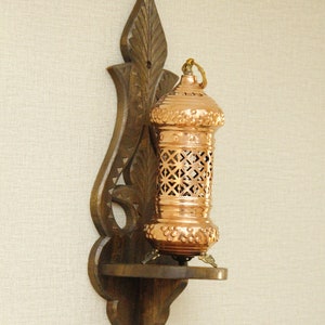 May include: A copper-colored lantern with a decorative, openwork design, mounted on a dark brown wooden wall sconce. The sconce has an ornate, carved design. The lantern has a small shelf and is designed for candles or small lights.