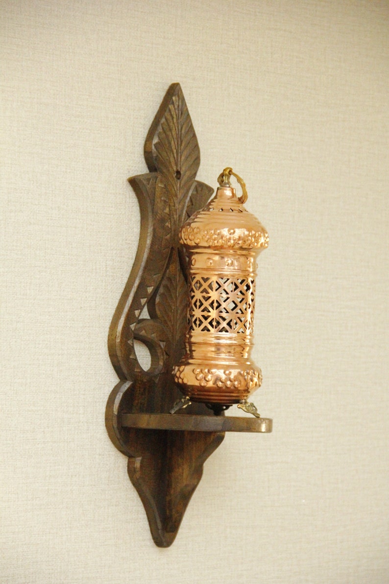 May include: A copper-colored lantern with a decorative, openwork design, mounted on a dark brown wooden wall sconce. The sconce has an ornate, carved design. The lantern has a small shelf and is designed for candles or small lights.