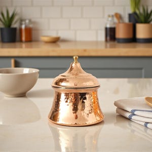 May include: A copper-colored, hammered-finish container with a lid and a small gold-colored knob. The container has a bulbous shape and sits on a white countertop. The background includes a kitchen setting with plants and other kitchenware.