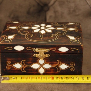 May include: A small wooden box with a hinged lid and a brass latch. The box is decorated with intricate gold and white floral patterns. The box is approximately 14 inches long.