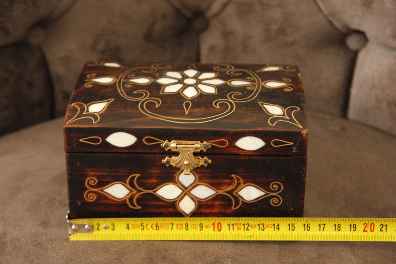 May include: A small wooden box with a hinged lid and a brass latch. The box is decorated with intricate gold and white floral patterns. The box is approximately 14 inches long.