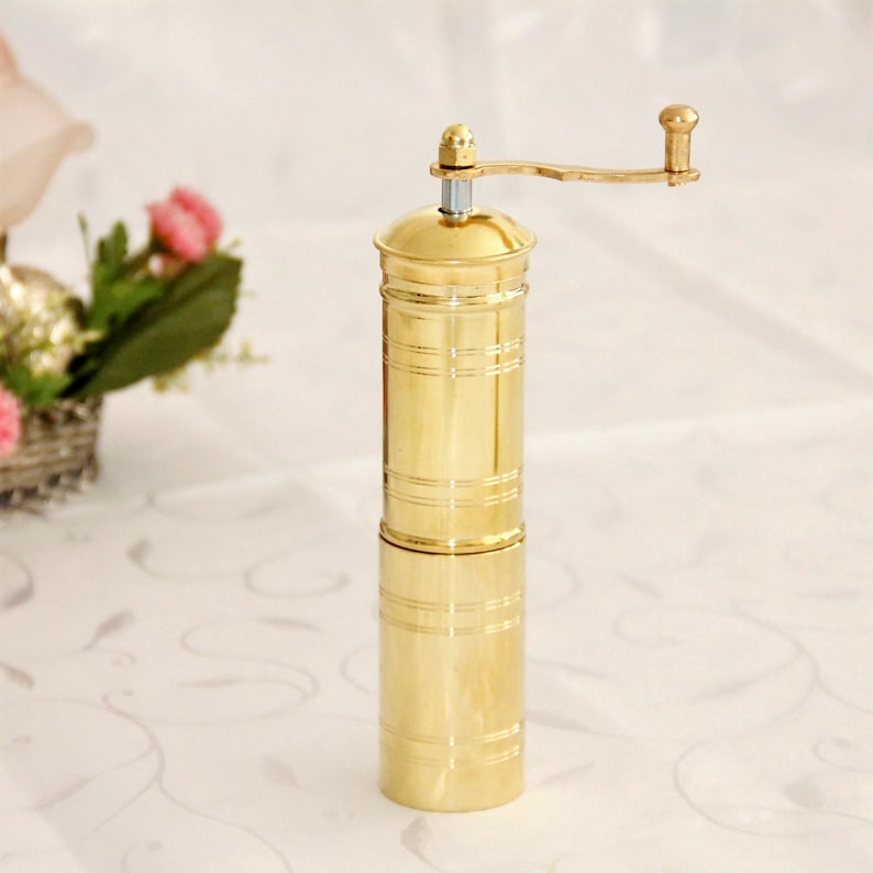 May include: A gold-colored manual coffee grinder with a crank handle. The grinder is made of metal and has a cylindrical shape.