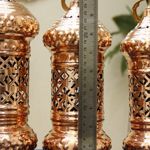 May include: Three copper-colored decorative lanterns with intricate cut-out designs. Each lantern is approximately 10 inches tall, with a handle at the top and ornate base. The lanterns are displayed on a blue surface.