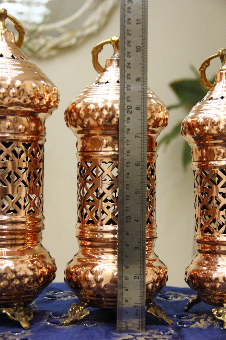 May include: Three copper-colored decorative lanterns with intricate cut-out designs. Each lantern is approximately 10 inches tall, with a handle at the top and ornate base. The lanterns are displayed on a blue surface.