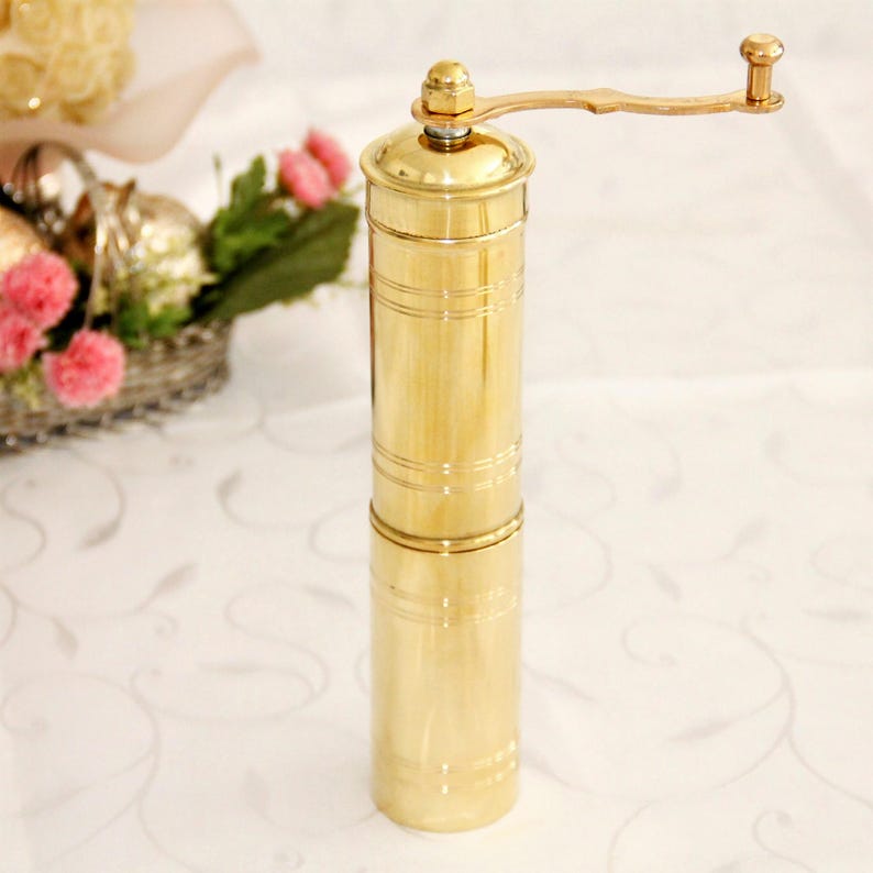 May include: A gold-colored manual pepper grinder with a crank handle. The grinder is made of metal and has a cylindrical shape.