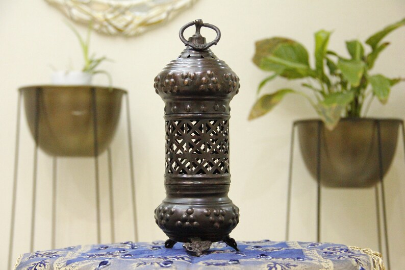 May include: A dark bronze metal lantern with a decorative design. The lantern has a cylindrical shape with a lattice pattern in the center and a ring at the top. The lantern is approximately 12 inches tall.