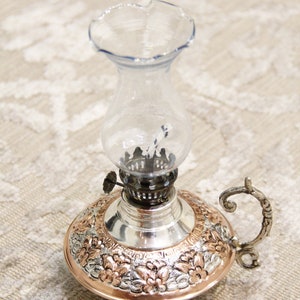 Pode incluir: Uma l&acirc;mpada de &oacute;leo de prata e cobre com uma chamin&eacute; de vidro e uma al&ccedil;a decorativa. A l&acirc;mpada tem um design floral.