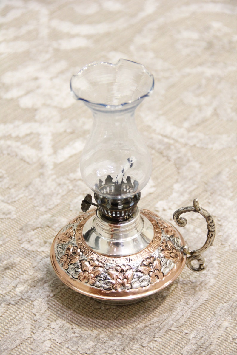 Pode incluir: Uma l&acirc;mpada de &oacute;leo de prata e cobre com uma chamin&eacute; de vidro e uma al&ccedil;a decorativa. A l&acirc;mpada tem um design floral.