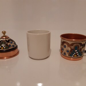Pode incluir: Um conjunto de tr&ecirc;s ch&aacute;venas, uma ch&aacute;vena de cer&acirc;mica branca e duas ch&aacute;venas de cobre com desenhos florais intrincados. As ch&aacute;venas de cobre t&ecirc;m uma pega preta e uma borda dourada.