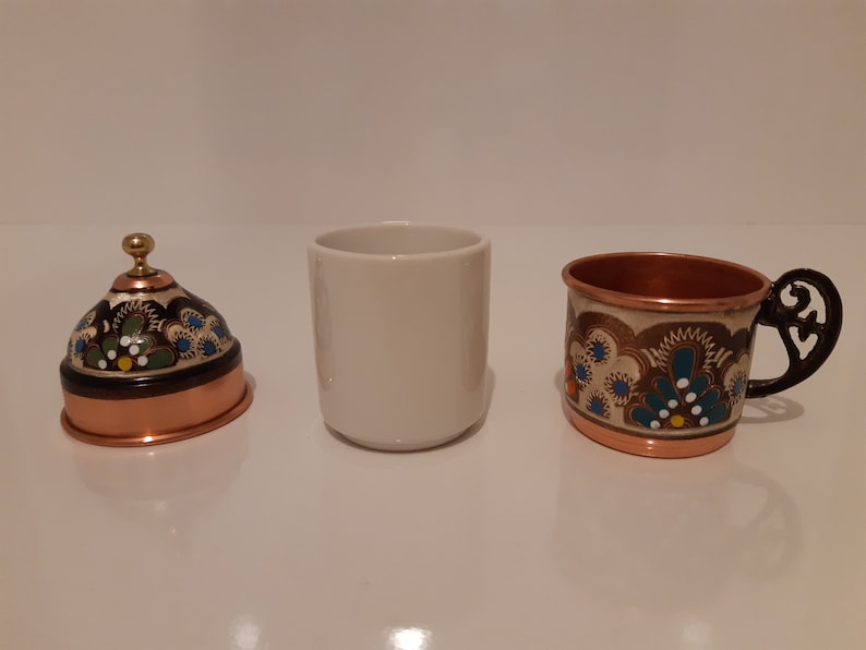 Pode incluir: Um conjunto de tr&ecirc;s ch&aacute;venas, uma ch&aacute;vena de cer&acirc;mica branca e duas ch&aacute;venas de cobre com desenhos florais intrincados. As ch&aacute;venas de cobre t&ecirc;m uma pega preta e uma borda dourada.