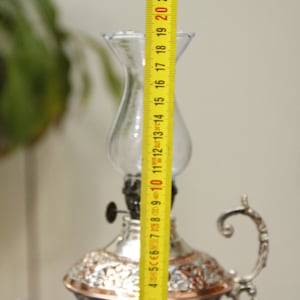 Pode incluir: Uma l&acirc;mpada de &oacute;leo de prata e cobre com uma chamin&eacute; de vidro transparente. A l&acirc;mpada tem 50,8 cm de altura e apresenta desenhos florais intrincados.
