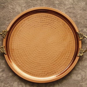 May include: A round copper serving tray with hammered texture and two ornate brass handles. The handles are shaped like grapevines with leaves.