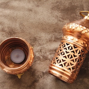 May include: A copper-colored candle lantern with a decorative geometric pattern and a small round handle. Beside it is a matching candle holder with a lit tealight candle inside. The items are on a brown surface.