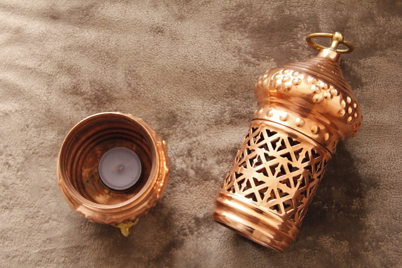 May include: A copper-colored candle lantern with a decorative geometric pattern and a small round handle. Beside it is a matching candle holder with a lit tealight candle inside. The items are on a brown surface.
