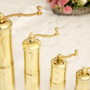 May include: Four gold-colored manual pepper mills of varying sizes. The mills have a crank handle and a cylindrical body with a decorative pattern.