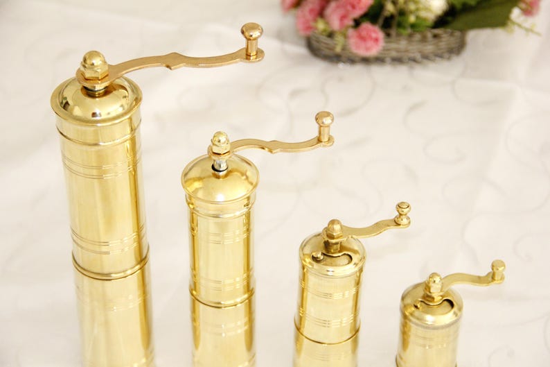 May include: Four gold-colored manual pepper mills of varying sizes. The mills have a crank handle and a cylindrical body with a decorative pattern.