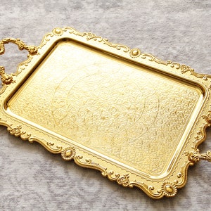 May include: A gold-colored serving tray with ornate detailing and two handles. The tray has a rectangular shape and a raised rim.