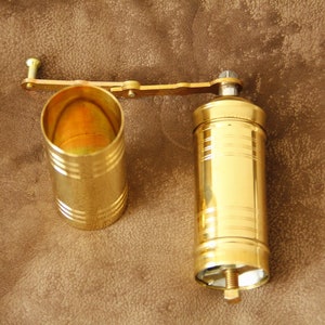May include: A gold metal coffee grinder with a crank handle. The grinder has a cylindrical shape and is made of two parts: a top section with a crank and a bottom section that holds the coffee beans.