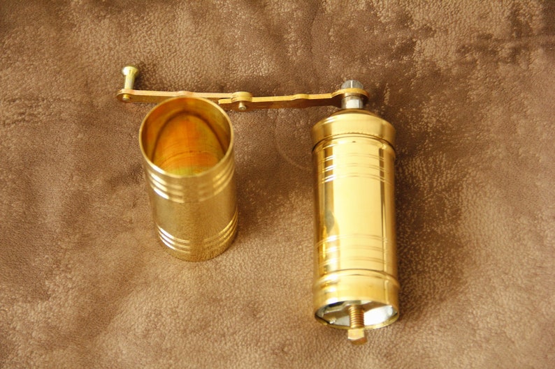 May include: A gold metal coffee grinder with a crank handle. The grinder has a cylindrical shape and is made of two parts: a top section with a crank and a bottom section that holds the coffee beans.