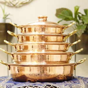 May include: A set of five copper cookware pieces with gold handles. The cookware is stacked in a pyramid shape and is sitting on a blue and gold patterned surface.