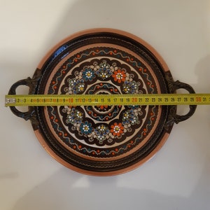 Pode incluir: Uma bandeja redonda de servir de cobre com uma borda marrom e duas al&ccedil;as. A bandeja &eacute; decorada com um padr&atilde;o floral colorido em tons de azul, verde, laranja e branco.