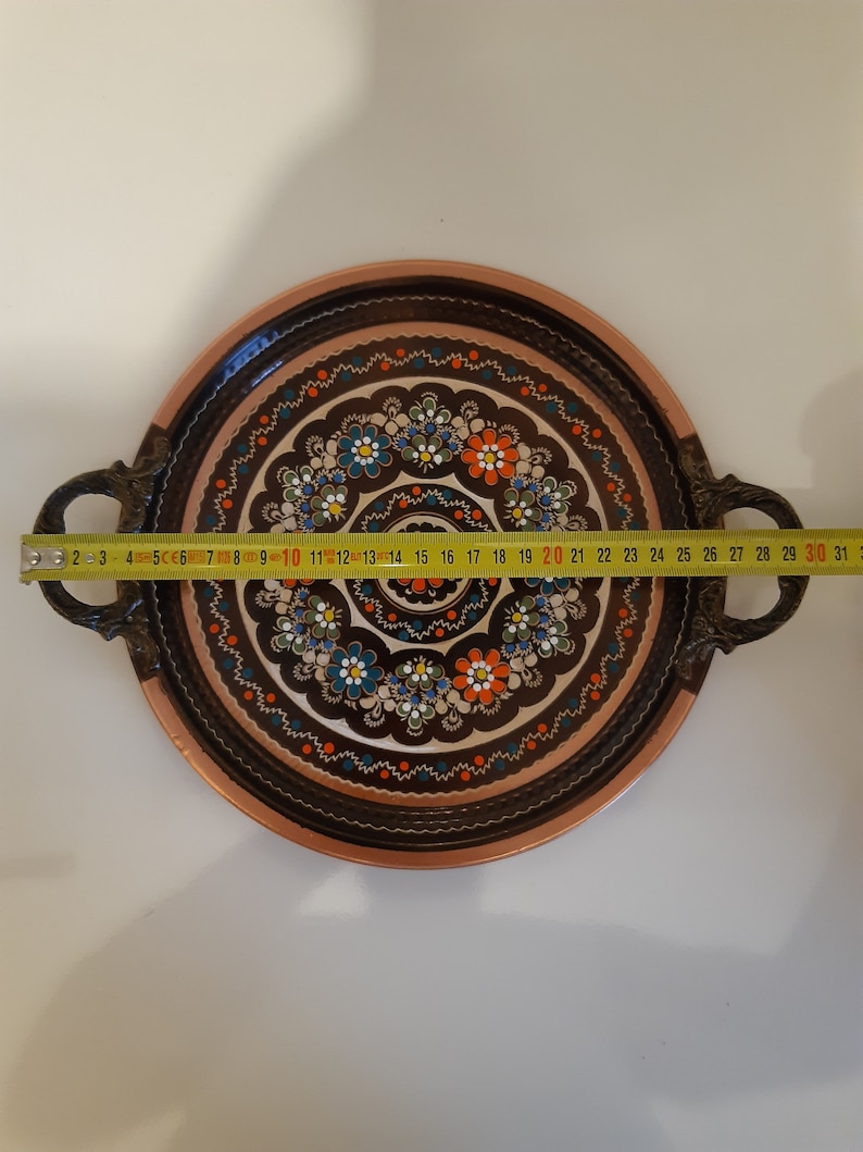 Pode incluir: Uma bandeja redonda de servir de cobre com uma borda marrom e duas al&ccedil;as. A bandeja &eacute; decorada com um padr&atilde;o floral colorido em tons de azul, verde, laranja e branco.