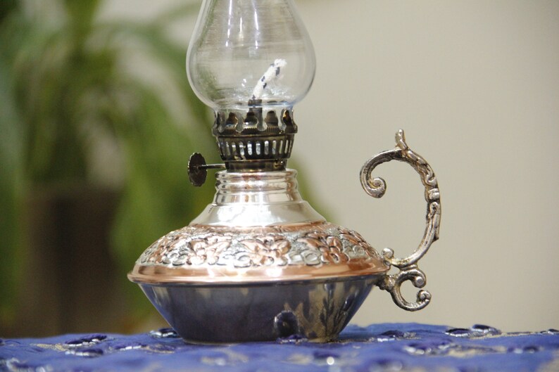 Pode incluir: Uma l&acirc;mpada de &oacute;leo de prata e cobre com uma chamin&eacute; de vidro e uma al&ccedil;a decorativa. A l&acirc;mpada tem um design ornamentado e &eacute; perfeita para adicionar um toque de charme vintage a qualquer casa.