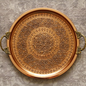 May include: A round copper serving tray with ornate embossed designs and two decorative handles.