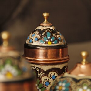 Pode incluir: Conjunto de quatro recipientes de cobre com desenhos pintados ornamentados em tons de azul, verde, branco e dourado. Os recipientes t&ecirc;m tampas abobadadas com remates.