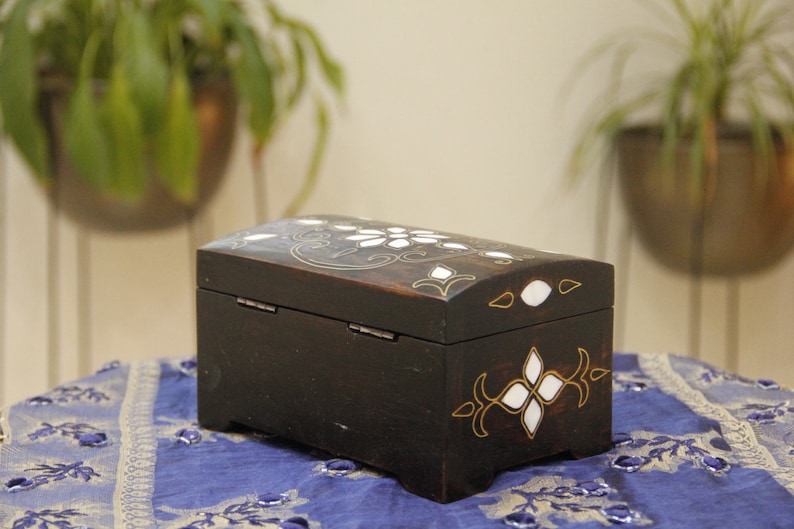 May include: A small, black wooden box with intricate gold and white inlay designs. The box has a hinged lid and is decorated with floral patterns.
