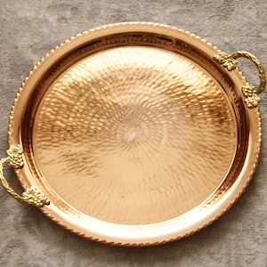 May include: A round copper serving tray with a hammered texture and two ornate gold handles. The tray has a beaded edge.