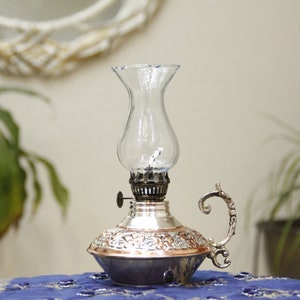 Pode incluir: Uma l&acirc;mpada de &oacute;leo de prata e cobre com uma chamin&eacute; de vidro transparente. A l&acirc;mpada tem um design ornamentado e uma al&ccedil;a.