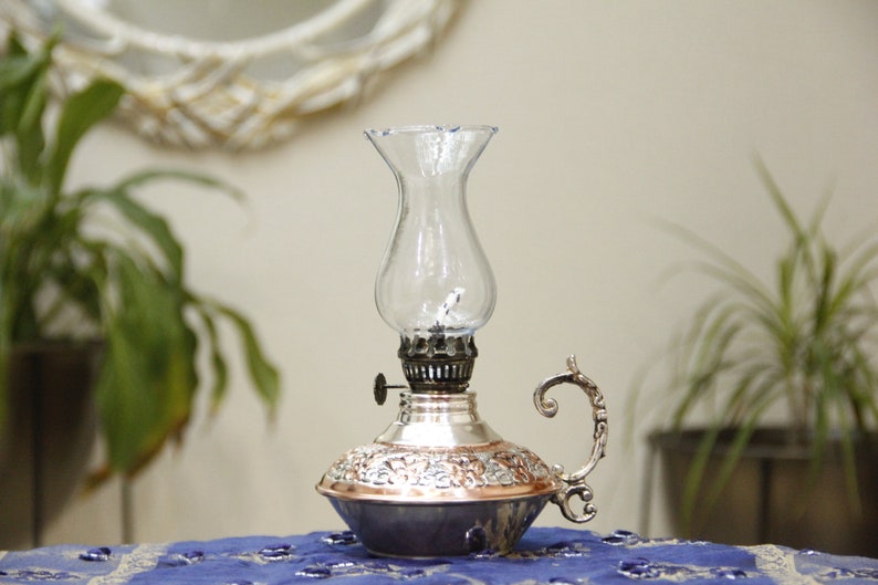Pode incluir: Uma l&acirc;mpada de &oacute;leo de prata e cobre com uma chamin&eacute; de vidro transparente. A l&acirc;mpada tem um design ornamentado e uma al&ccedil;a.