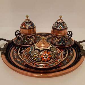 Pode incluir: Uma bandeja de servir de cobre com duas x&iacute;caras de caf&eacute; turco ornamentadas e seus pires, e um pequeno a&ccedil;ucareiro de cobre com tampa. A bandeja e as x&iacute;caras s&atilde;o decoradas com desenhos florais pintados &agrave; m&atilde;o em tons de azul, verde, laranja e branco.
