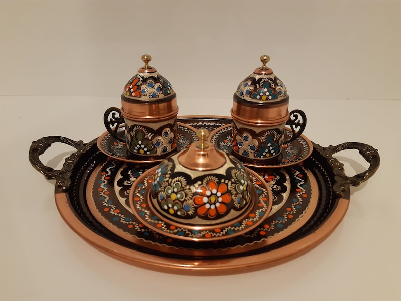 Pode incluir: Uma bandeja de servir de cobre com duas x&iacute;caras de caf&eacute; turco ornamentadas e seus pires, e um pequeno a&ccedil;ucareiro de cobre com tampa. A bandeja e as x&iacute;caras s&atilde;o decoradas com desenhos florais pintados &agrave; m&atilde;o em tons de azul, verde, laranja e branco.