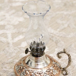 Pode incluir: Uma l&acirc;mpada de &oacute;leo de cobre e prata com uma chamin&eacute; de vidro transparente e um design floral decorativo. A l&acirc;mpada tem uma al&ccedil;a e um pavio.