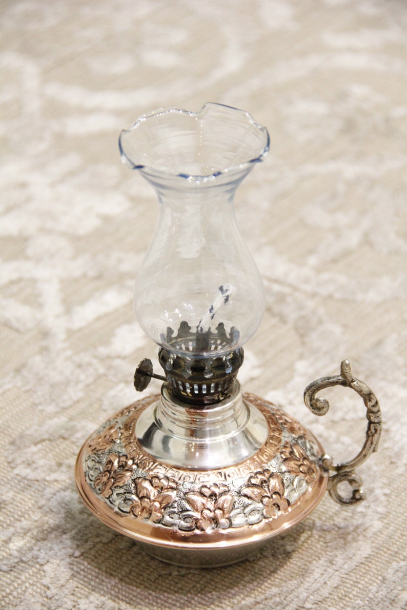 Pode incluir: Uma l&acirc;mpada de &oacute;leo de cobre e prata com uma chamin&eacute; de vidro transparente e um design floral decorativo. A l&acirc;mpada tem uma al&ccedil;a e um pavio.