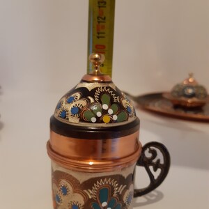 Pode incluir: Uma pequena cafeteira de cobre com tampa e al&ccedil;a. A cafeteira &eacute; decorada com um padr&atilde;o floral em azul, verde e branco.