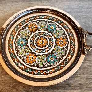Pode incluir: Uma bandeja redonda de servir de cobre com duas al&ccedil;as. A bandeja tem um fundo preto com um desenho floral colorido em tons de laranja, verde e azul.