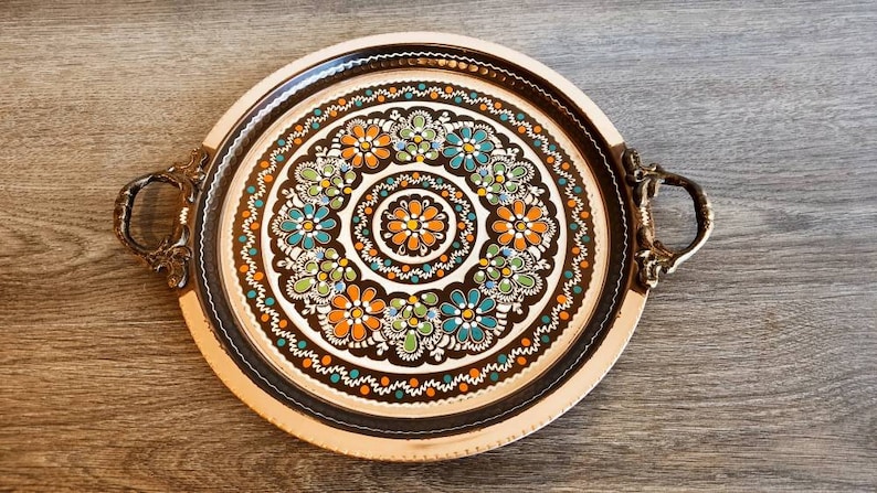 Pode incluir: Uma bandeja redonda de servir de cobre com duas al&ccedil;as. A bandeja tem um fundo preto com um desenho floral colorido em tons de laranja, verde e azul.