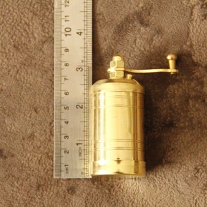 May include: A gold-colored metal pepper grinder with a crank handle. The grinder is about 2 inches tall and is sitting on a brown surface.