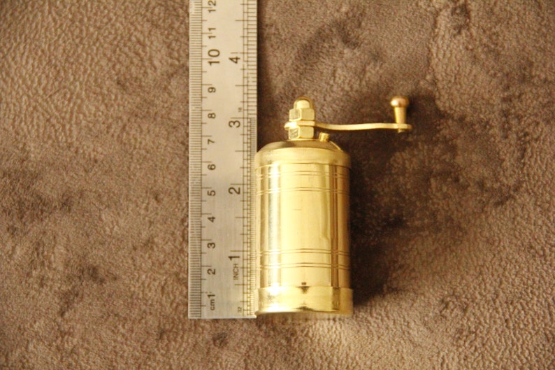 May include: A gold-colored metal pepper grinder with a crank handle. The grinder is about 2 inches tall and is sitting on a brown surface.