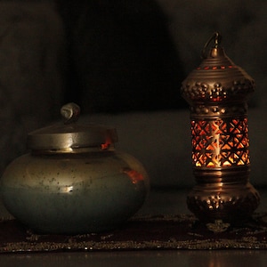 May include: A copper-colored lantern with a geometric pattern and a small, round, lidded jar. The lantern is lit from within, casting a warm glow. The jar is a muted teal color with a silver lid.